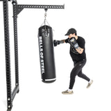 A person in a black outfit and cap boxes a black Heavy Bag Set from Bells of Steel, hanging from a sturdy mount. With boxing gloves and checkered shoes against a white background, they capture the intensity of Combat Sports.