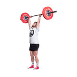 A man in a gray t-shirt, black shorts, and red sneakers lifts a barbell with Bells of Steel Boltless LB Competition Bumper Plates overhead on a white background.