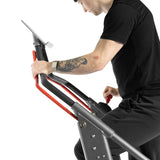 Wearing black workout clothes and a smartwatch, a tattooed user grips the red and black handles of the Bells of Steel Sisyphean Stepper – Manual Stair Climber, exercising on this cardio machine against a white background.