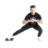 A man wearing a black Bells of Steel T-shirt and pants does a lateral lunge on a Glute Ham Slider, focusing on hamstrings. He balances with hands in front of his chest and sports white sneakers on a plain white background.