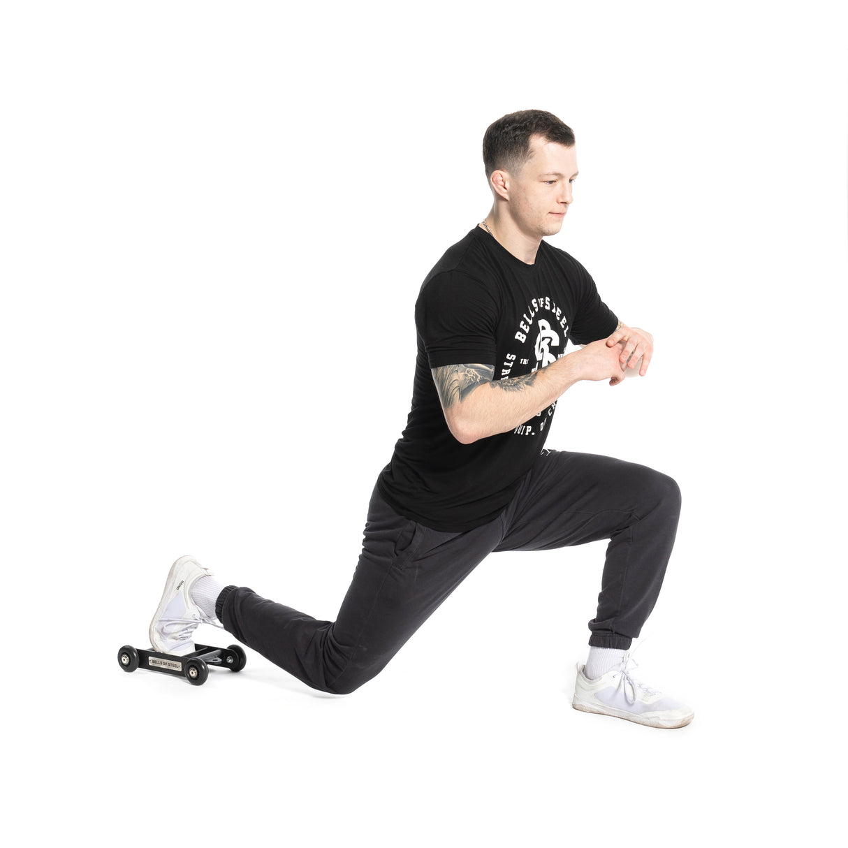 A person wearing a black outfit performs a lunge stretch on a Bells of Steel Glute Ham Slider, with their back knee bent and arms extended forward. Their light shoes contrast the minimalist white background, highlighting hamstring movements.