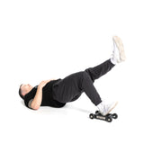 Lying on their back in black pants, a black shirt, and white sneakers against a white background, a person performs hamstring movements using the Bells of Steel Glute Ham Slider with heavy-duty wheels, keeping one leg bent and the other extended upward while maintaining perfect form.