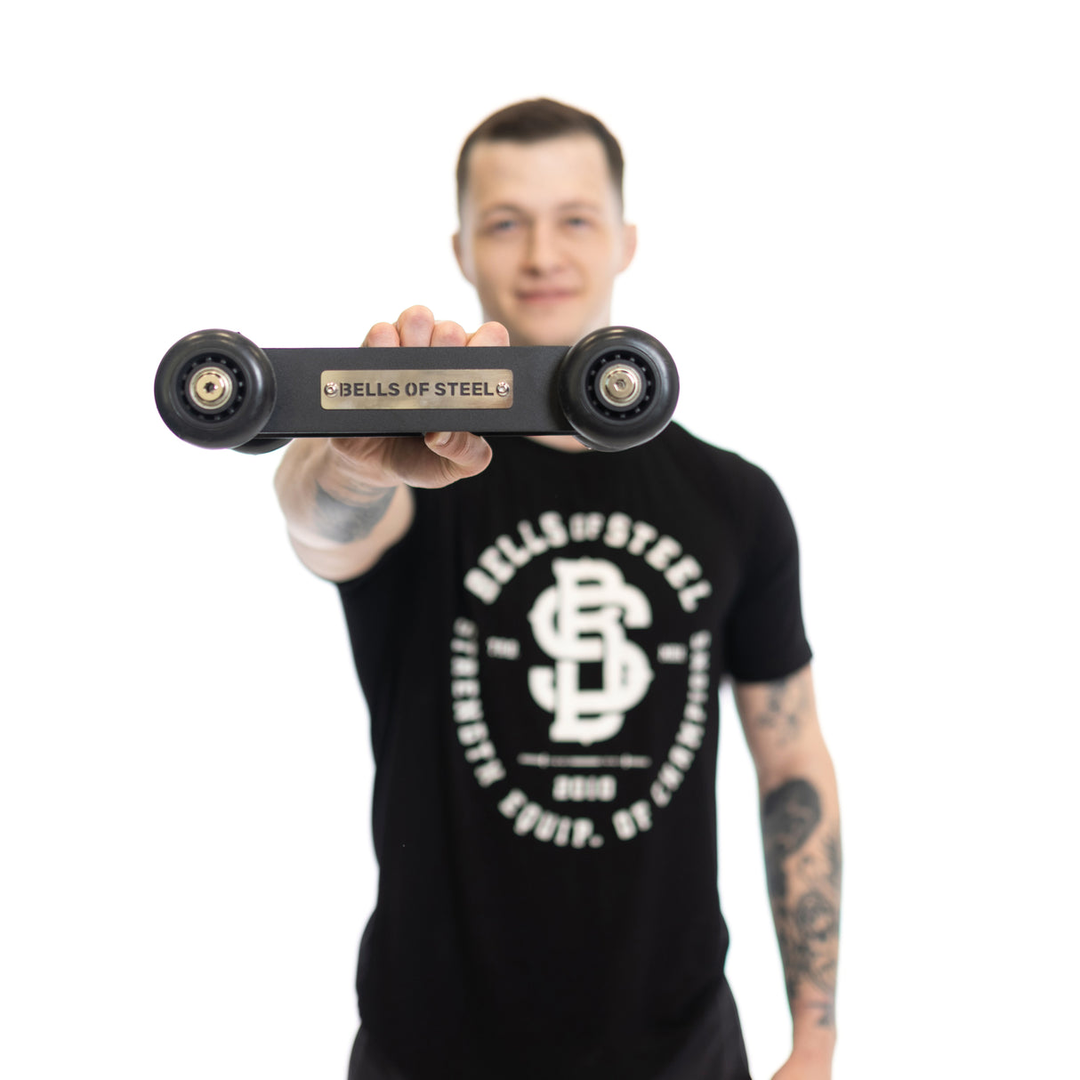 A person in a black Bells of Steel t-shirt presents a skateboard truck featuring the brand's engraving. The crisp white backdrop highlights their pose, suggesting strength reminiscent of using the Glute Ham Slider.