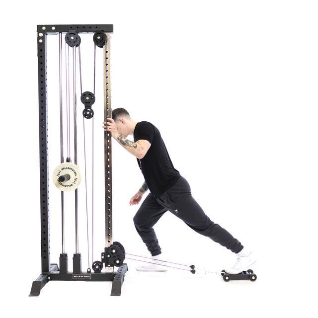 Dressed in black athletic wear, an individual exercises on a cable machine, using their leg muscles and posterior chain. They lean forward with one foot on the Bells of Steel Glute Ham Slider connected to a cable, against a white background that accentuates the dynamic movement.