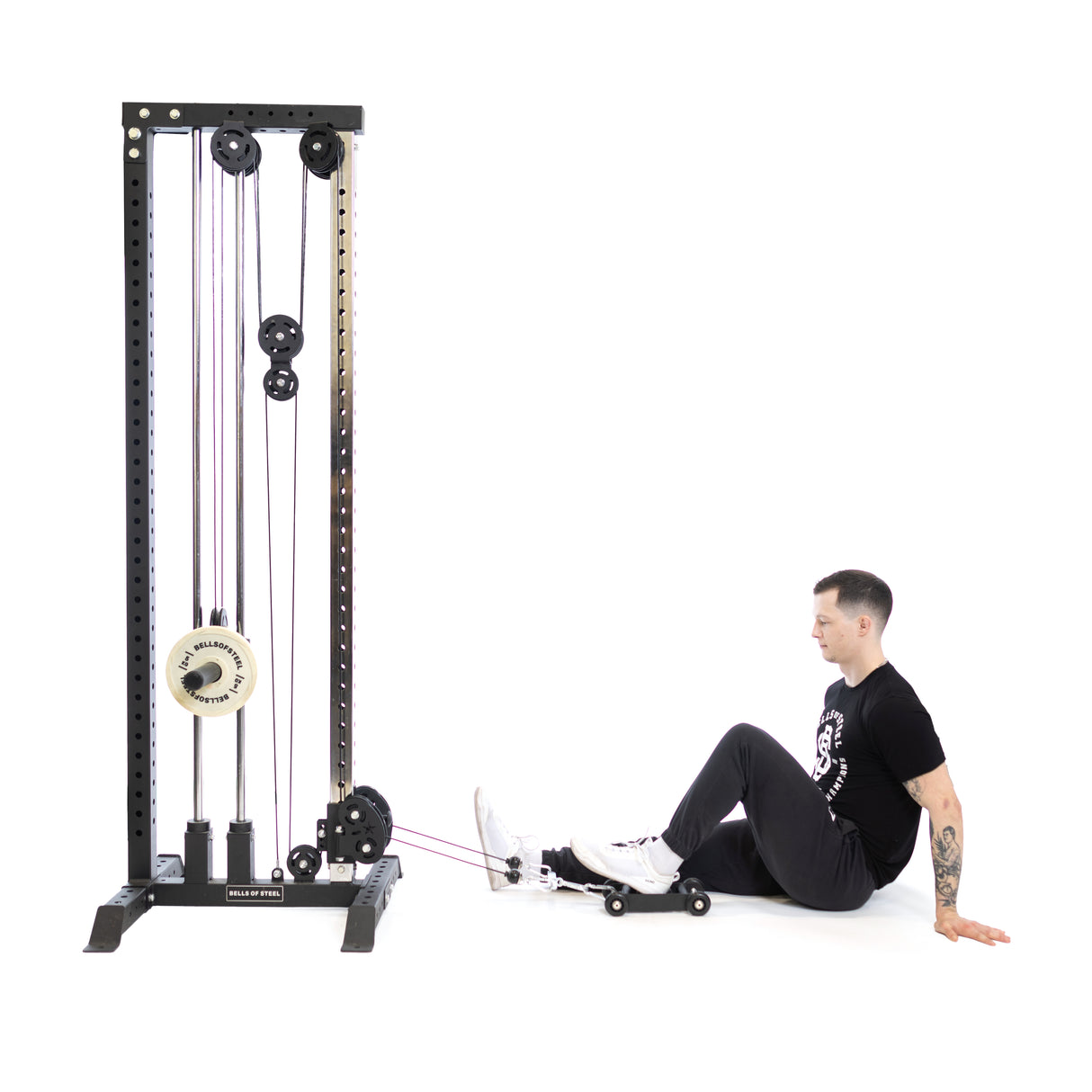 A tattooed individual is seated on the floor beside a cable machine, using Bells of Steel's Glute Ham Slider with resistance bands. Wearing a black T-shirt and dark pants, they are intensely strengthening their posterior chain through a seated leg exercise.