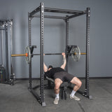 Wearing black shorts and a T-shirt, a person bench presses with a barbell inside a power rack featuring the Bells of Steel Monolift Rack Attachment, creating a more space-saving gym setup.