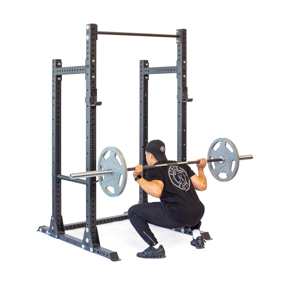 A person in workout attire performs a squat with a barbell on the Hydra Half Rack Builder (3" x 3", ⅝" Holes) from Bells of Steel. Wearing a cap, they showcase strength and focus against the white backdrop of their home gym.