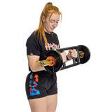 A red-haired woman in a ponytail lifts a heavy barbell fitted with Bells of Steel Magnetic Clamp Collars. She wears a black T-shirt with colorful text and black shorts featuring blue and red details, standing against a white background.