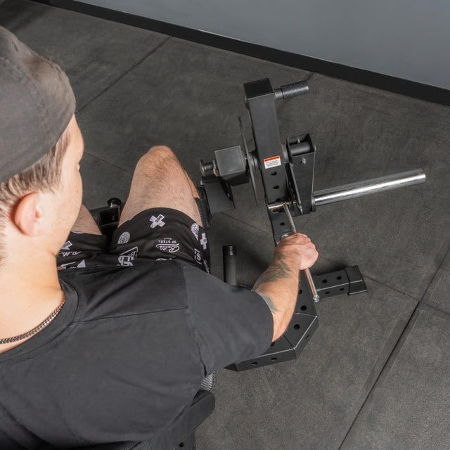A person in a black shirt, black cap, and patterned shorts adjusts the Bells of Steel USA Legacy Leg Extension / Hamstring Curl Machine – Plate Loaded in a home gym with gray flooring. The photo is taken from above and behind.