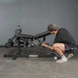 A person in a cap and shorts adjusts the settings on the Bells of Steel USA Legacy Leg Extension / Hamstring Curl Machine - Plate Loaded, ideal for home gyms, in a gym with gray walls and floor.