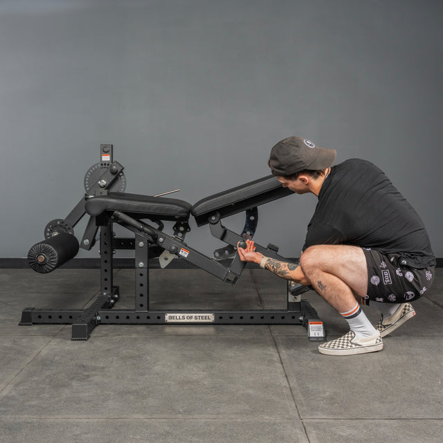 A person in a cap and shorts adjusts the settings on the Bells of Steel USA Legacy Leg Extension / Hamstring Curl Machine - Plate Loaded, ideal for home gyms, in a gym with gray walls and floor.