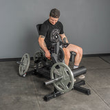 A man in a black t-shirt, shorts, and backward cap adjusts the Bells of Steel USA Legacy Leg Extension / Hamstring Curl Machine - Plate Loaded in a gym with gray walls and flooring.