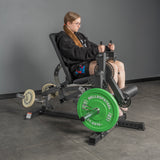 A woman with long hair, glasses, and a black sweatshirt uses the Bells of Steel USA Legacy Leg Extension / Hamstring Curl Machine—plate loaded with green and white weights—in a gray-walled, concrete-floored home gym.