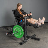 A woman with long hair and glasses uses a Bells of Steel USA Legacy Leg Extension / Hamstring Curl Machine - Plate Loaded in a gym, wearing a black hoodie, shorts, and pink sneakers. The machine has green weight plates on a gray floor and walls.