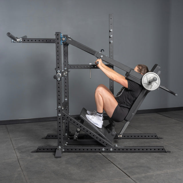 Wearing gym attire, a person uses the Bells of Steel USA Pandemonium Squat – a 3-in-1 Pendulum Squat / Calf Press / Viking Press Machine – with weights, set in a minimalist gray-walled multifunctional home gym.