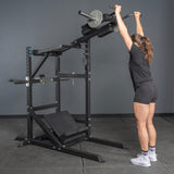 A woman in athletic wear stands before the Bells of Steel USA Pandemonium Squat – 3-in-1 Pendulum Squat / Calf Press / Viking Press Machine, reaching up to grip the handles as she prepares for an overhead exercise.