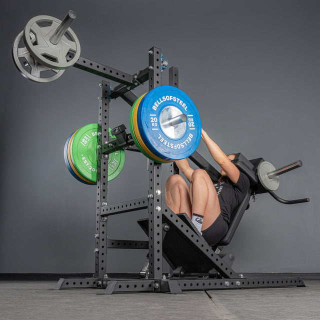 A person uses the Bells of Steel USA Pandemonium Squat – 3-in-1 Pendulum Squat / Calf Press / Viking Press Machine, loaded with colorful weight plates, in a gym with gray walls and a concrete floor, pushing weights upward with their legs.