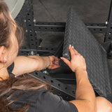 A person with long brown hair adjusts the textured metal footplate on the Bells of Steel USA Pandemonium Squat – a 3-in-1 Pendulum Squat / Calf Press / Viking Press Machine, gripping and moving the angled plate indoors on a dark floor.
