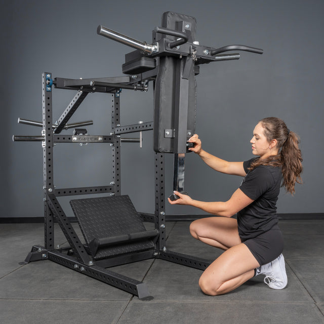 A woman in athletic wear kneels beside the Bells of Steel USA Pandemonium Squat – 3-in-1 Pendulum Squat / Calf Press / Viking Press Machine, adjusting a padded bar. The equipment is set against a gray wall on a concrete gym floor.