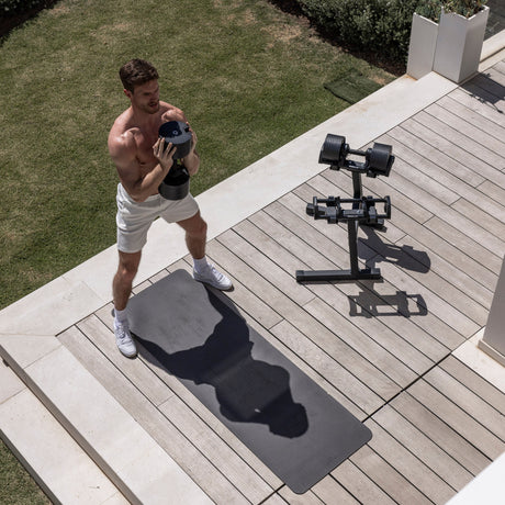 A man in white shorts and sneakers stands on an exercise mat outdoors, holding a dumbbell. Nearby, the Nuobell NÜOBELL Floor Stand stores adjustable dumbbells compactly amid grass and plants in the sunny background.