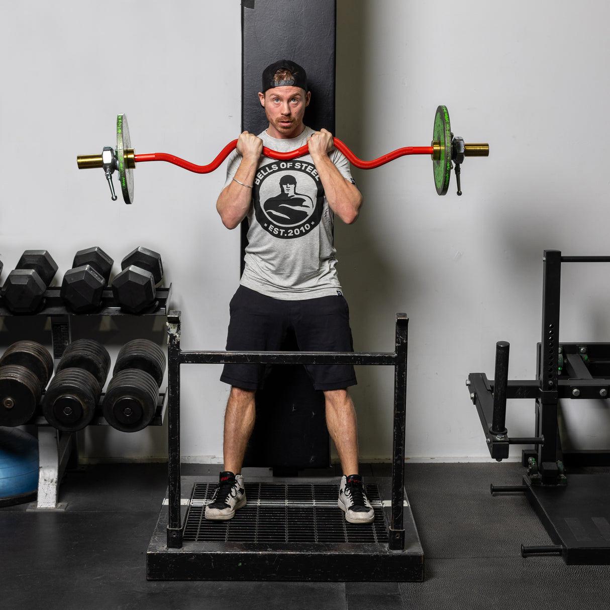 A man in a gray shirt, black shorts, and sneakers grips the Bells of Steel 100% RAW Competition Curl Bar with green weights at shoulder height in a power rack. Dumbbells sit on a rack to his left against the plain gym wall.