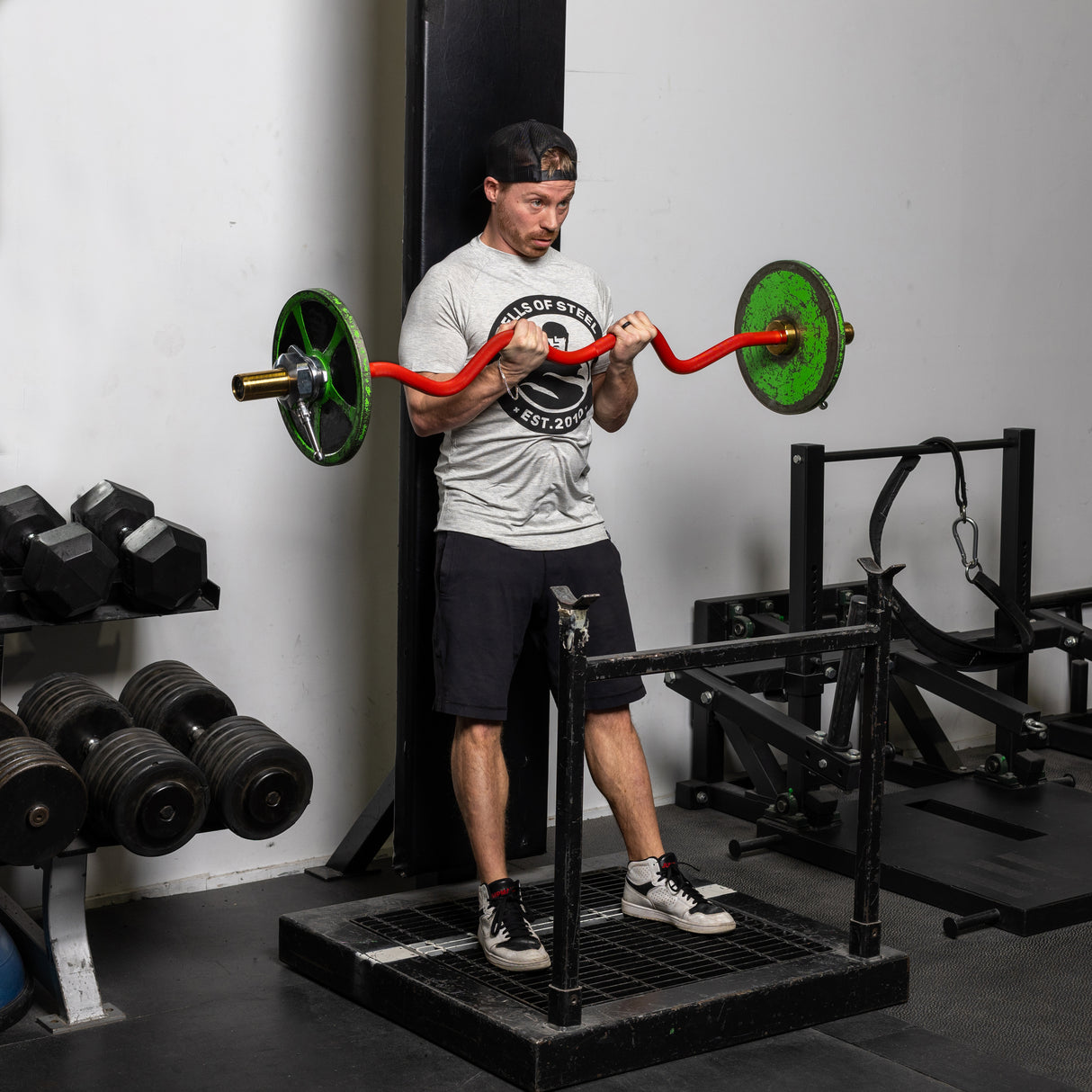 A man in a gym is doing bicep curls with a Bells of Steel 100% RAW Competition Curl Bar, featuring an aggressive knurl. He wears a backward cap, gray T-shirt, and black shorts. Dumbbells and gym equipment fill the background.