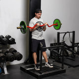 A man in a gym is doing bicep curls with a Bells of Steel 100% RAW Competition Curl Bar, featuring an aggressive knurl. He wears a backward cap, gray T-shirt, and black shorts. Dumbbells and gym equipment fill the background.