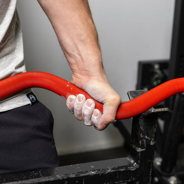 A hand grips a red, chalk-dusted 100% RAW Competition Curl Bar by Bells of Steel, against a backdrop of gym equipment. The person wears a light gray shirt and dark pants, highlighting dedication and focus.