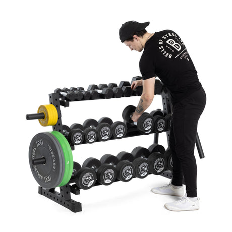 A person in a black outfit and cap arranges weights on Bells of Steel's Residential Dumbbell Rack, stocked with hex dumbbells and barbell plates. The minimalist setup creates the perfect home gym vibe against a plain white background.