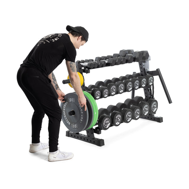 Dressed in black athletic wear, a person with a cap and white sneakers is lifting a weight plate from the Bells of Steel Residential Dumbbell Rack filled with hex dumbbells and weights in a gym setting.