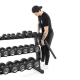 A person dressed in black adjusts a bar on the Bells of Steel Residential Dumbbell Rack, filled with various weights. The white background highlights their focus on organizing or securing the home gym equipment.