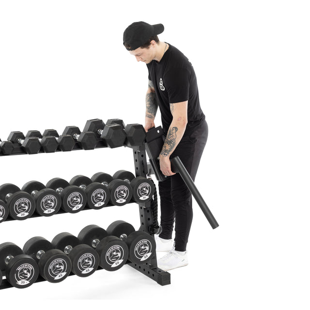 A person dressed in black adjusts a bar on the Bells of Steel Residential Dumbbell Rack, filled with various weights. The white background highlights their focus on organizing or securing the home gym equipment.