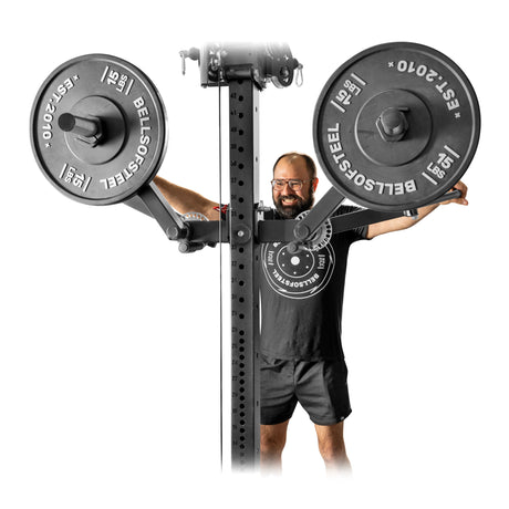 A person wearing glasses and a black t-shirt stands behind the Bells of Steel Shoulder Boulder/Chest Fly Attachment, smiling with arms outstretched near two “Bells of Steel” weight plates—a perfect space-saving choice for your next shoulder workout.