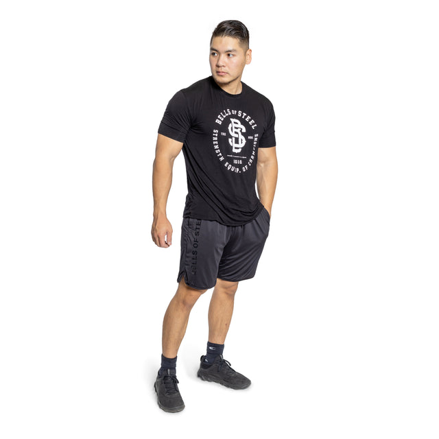 A man with short hair models stretchy, comfortable gym shorts from Bells of Steel against a plain white background. He wears a black t-shirt featuring white text and looks to the side to highlight the shorts' high-performance polyester material.