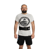 A bearded man with glasses wears a Bells of Steel t-shirt, black shorts, and the Bells of Steel Self Locking Belt, standing against a plain white background.