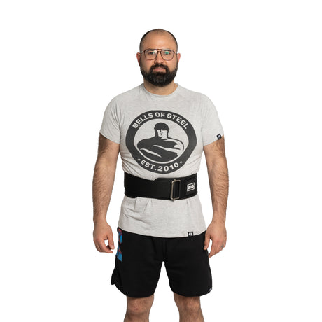 A bearded man with glasses wears a Bells of Steel t-shirt, black shorts, and the Bells of Steel Self Locking Belt, standing against a plain white background.