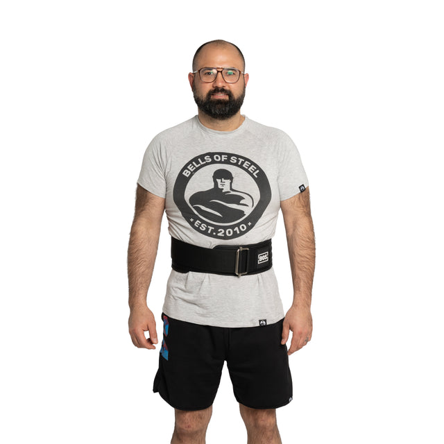 A bearded man with glasses wears a Bells of Steel t-shirt, black shorts, and the Bells of Steel Self Locking Belt, standing against a plain white background.