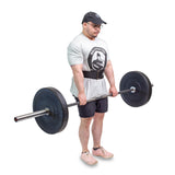 A person wearing a gray shirt, black shorts, cap, and glasses is using the Bells of Steel Self Locking Belt for back support while holding a loaded barbell, preparing to deadlift. The background is white.