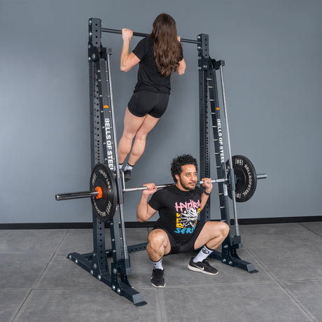 A woman does a pull-up on a Bells of Steel USA Smith Machine - Hydra (3" x 3", ⅝" Holes), while a man squats with a barbell beneath her in a gym featuring gray walls and tile flooring.