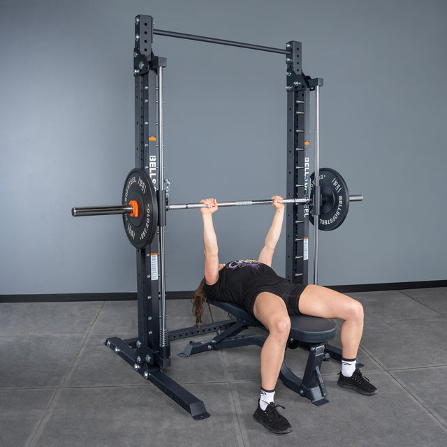 A person prepares to bench press on the Bells of Steel USA Smith Machine - Hydra (3" x 3", ⅝" holes) in a gym, gripping the barbell while lying on a bench next to a squat stand.