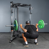Wearing athletic gear, an individual performs a barbell squat with green weight plates inside a Bells of Steel All-in-One Trainer, set in a home gym with gray walls and floor.