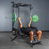 A person is bench pressing a barbell with green plates using the Bells of Steel All-in-One Trainer power rack in a gym with gray walls and black flooring.