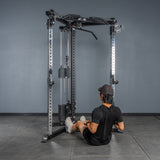 A person in a black shirt, shorts, and cap sits on a gym floor with tile flooring and a gray wall, holding handles and preparing for a seated row on the Bells of Steel All-in-One Trainer.