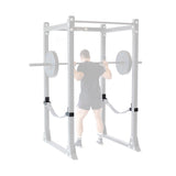 A person in gym attire performs a barbell squat in a Bells of Steel power rack, with faded weights and rack highlighting the Safety Strap Connectors, which stand out to emphasize training safety and focus on the lifter in full color.