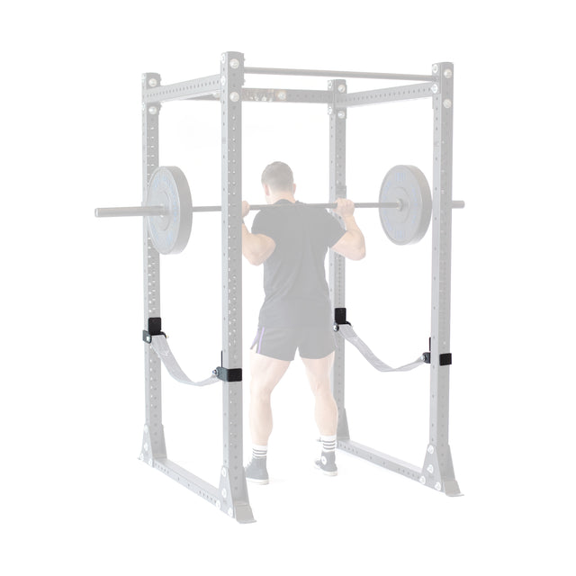 A person in gym attire performs a barbell squat in a Bells of Steel power rack, with faded weights and rack highlighting the Safety Strap Connectors, which stand out to emphasize training safety and focus on the lifter in full color.