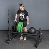Wearing athletic gear, a person loads weight plates onto the Bells of Steel USA Rack Junctions - 3" x 3" in a gym. A black medicine ball and an empty barbell rest near the gray wall and floor.