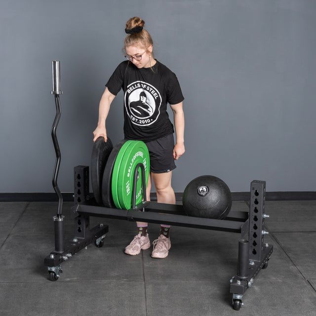 Wearing athletic gear, a person loads weight plates onto the Bells of Steel USA Rack Junctions - 3" x 3" in a gym. A black medicine ball and an empty barbell rest near the gray wall and floor.