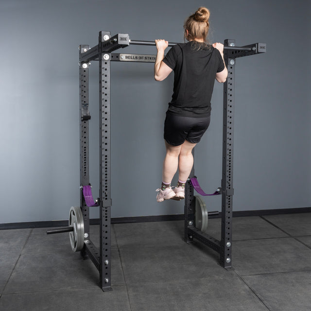 A person in black athletic wear does a pull-up on the Bells of Steel USA Rack Junctions - 3" x 3", a customizable rack setup with side weights, in a gym with gray walls and flooring.