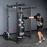 A man in black athletic wear and gray shoes punches a black BOS punching bag hanging from a Bells of Steel USA Rack Junctions - 3" x 3" setup in a home gym, with weight plates stacked on the rack.