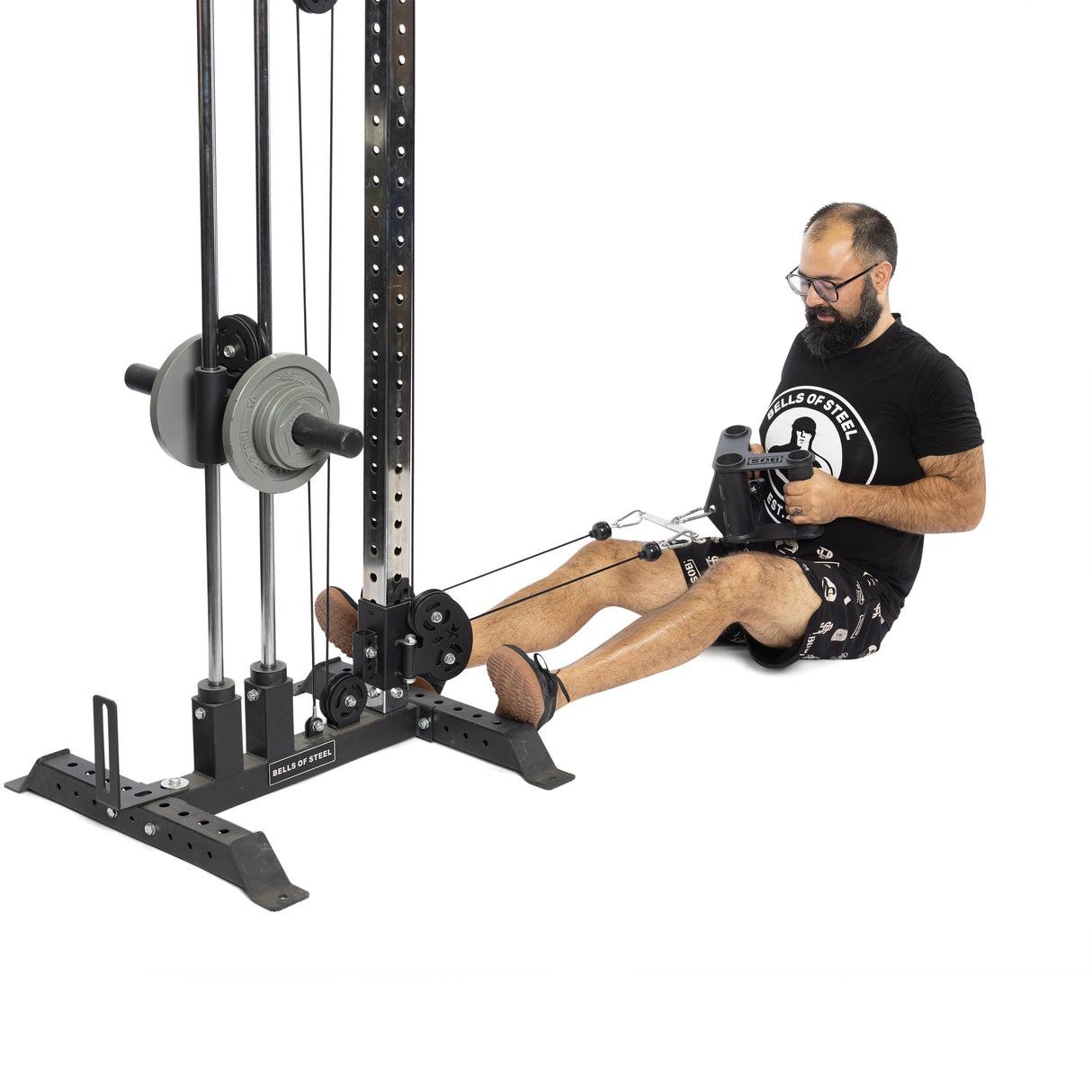 A man with a beard in a black t-shirt, graphic print shorts, and shoes, sits using the Bells of Steel Fat Bar - T-Bar Row Cable Attachment to enhance grip strength. Weight plates are attached for optimal resistance.